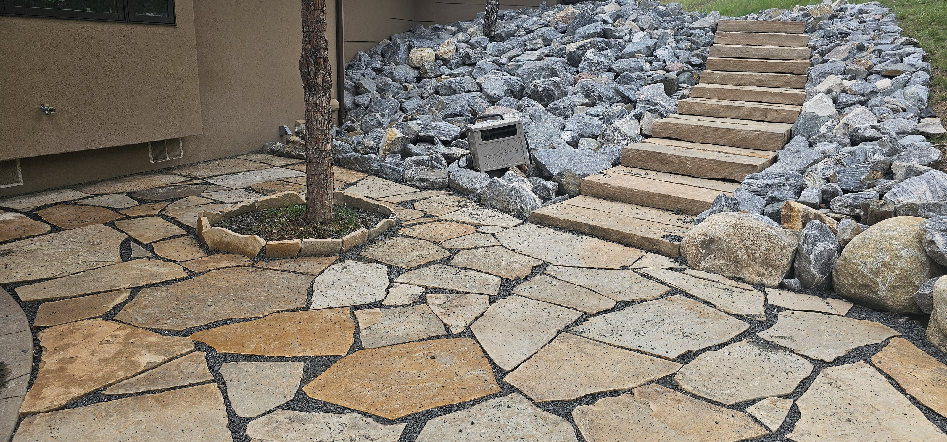 Flagstone patio, a tree planted in the paving, and stacked stone steps up a rocky slope.