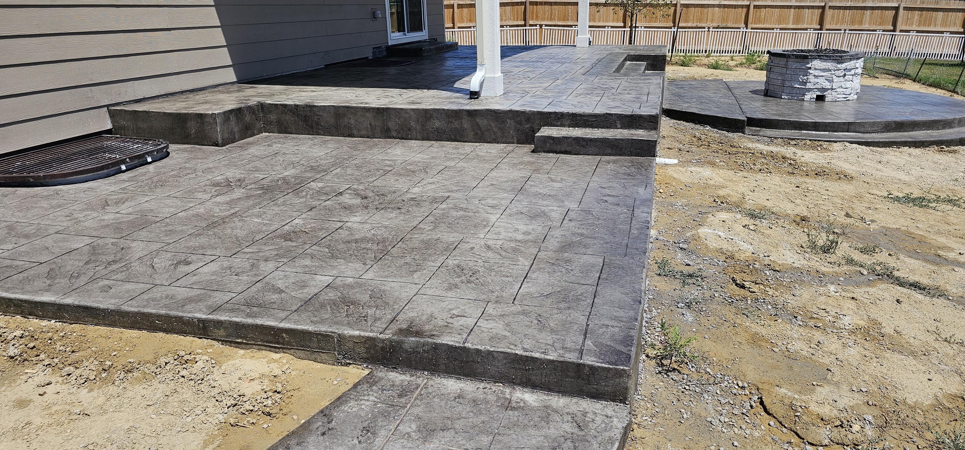 Dark gray stamped concrete patio with a lighter border and steps, landscaping unfinished beside it.