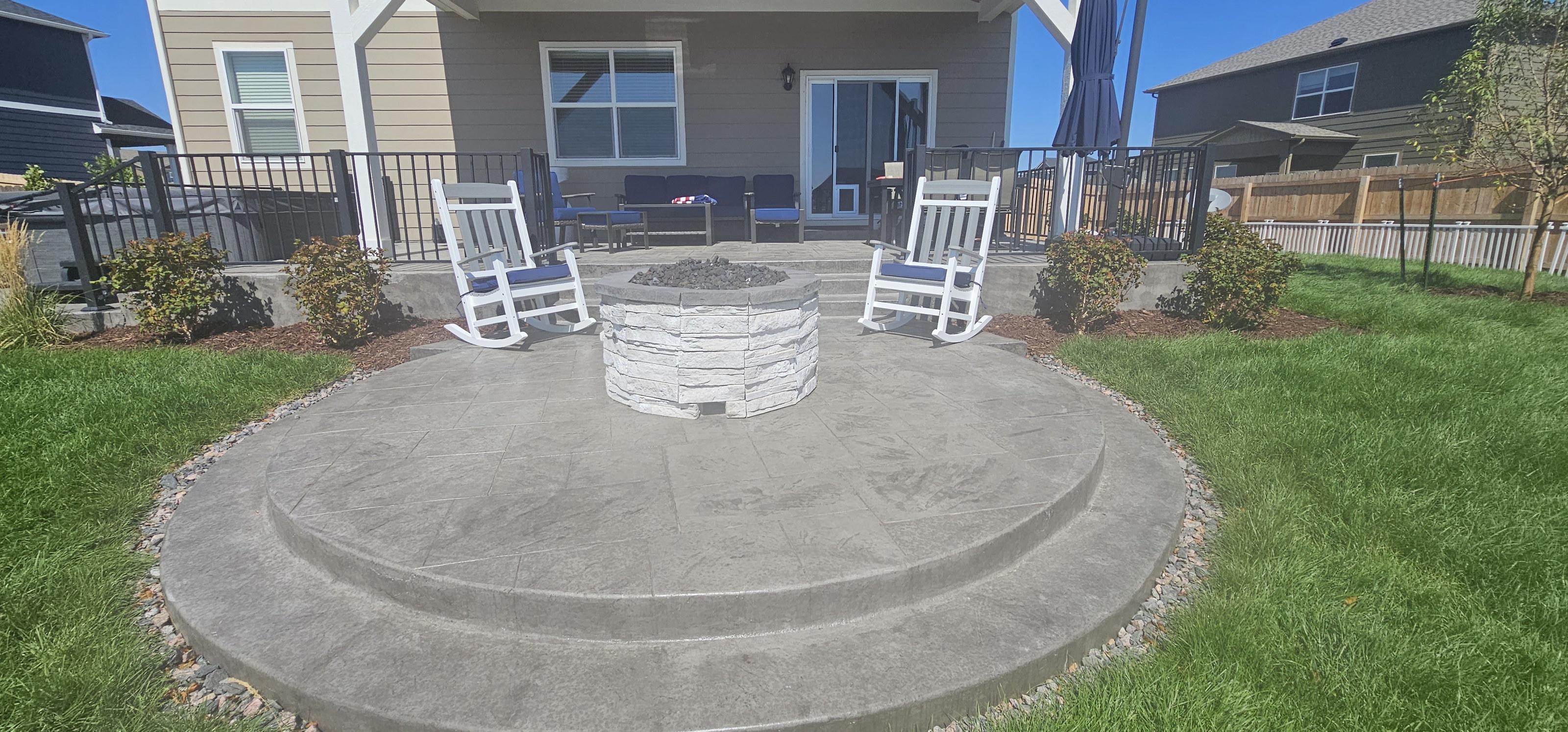 Circular stamped concrete patio with a fire pit, rocking chairs, and a fenced lawn.